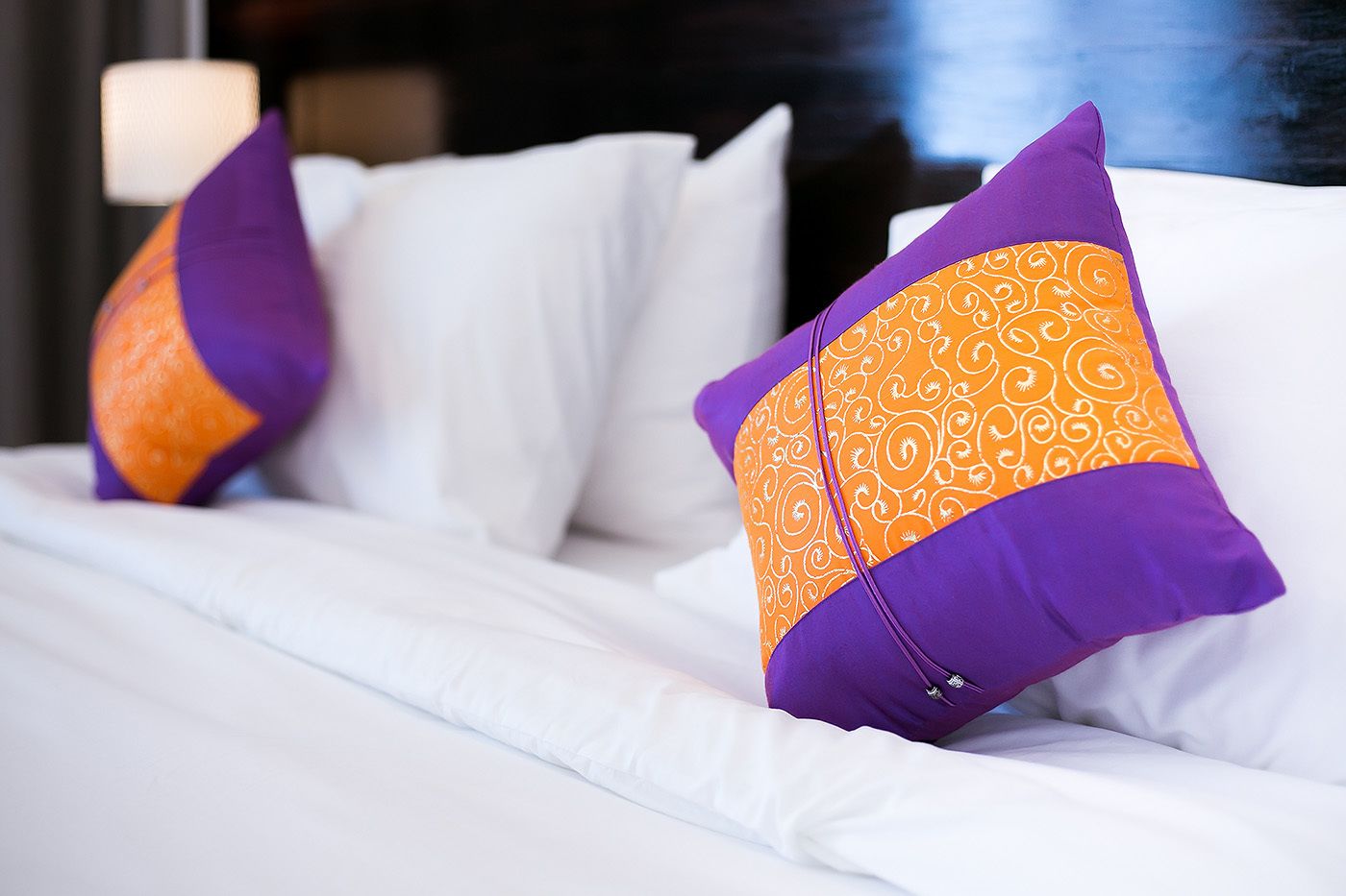 A bed with white bedding and two decorative pillows, one purple and orange, the other purple and white.