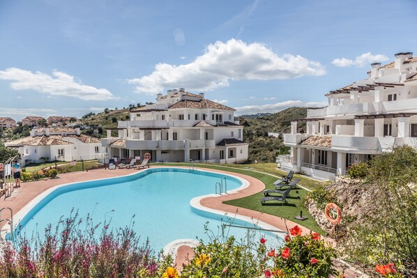 A swimming pool surrounded by white buildings with terracotta roofs, a clear sky, and a landscaped area with flowers and a lifebuoy.
