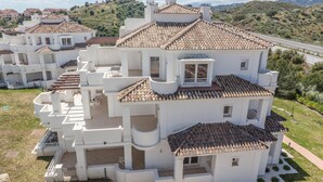 A white villa with multiple balconies and a tiled roof, surrounded by greenery and a road.