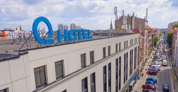 Un hotel con un letrero azul llamativo, una fila de coches aparcados a lo largo de la calle y un paisaje urbano al fondo.