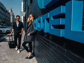Egy modern dizájnú, feltűnő hotelszign, egy kiépített járdaszakasz és egy parkoló autók által szegélyezett városi utca.