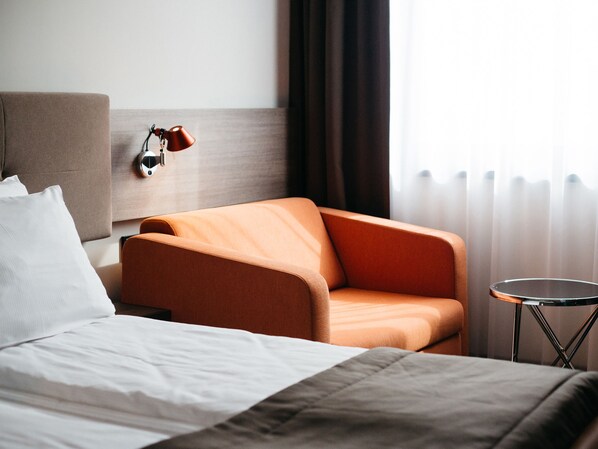 Una habitación de hotel con una cama, un sillón naranja, una mesita redonda y una lámpara de pared.