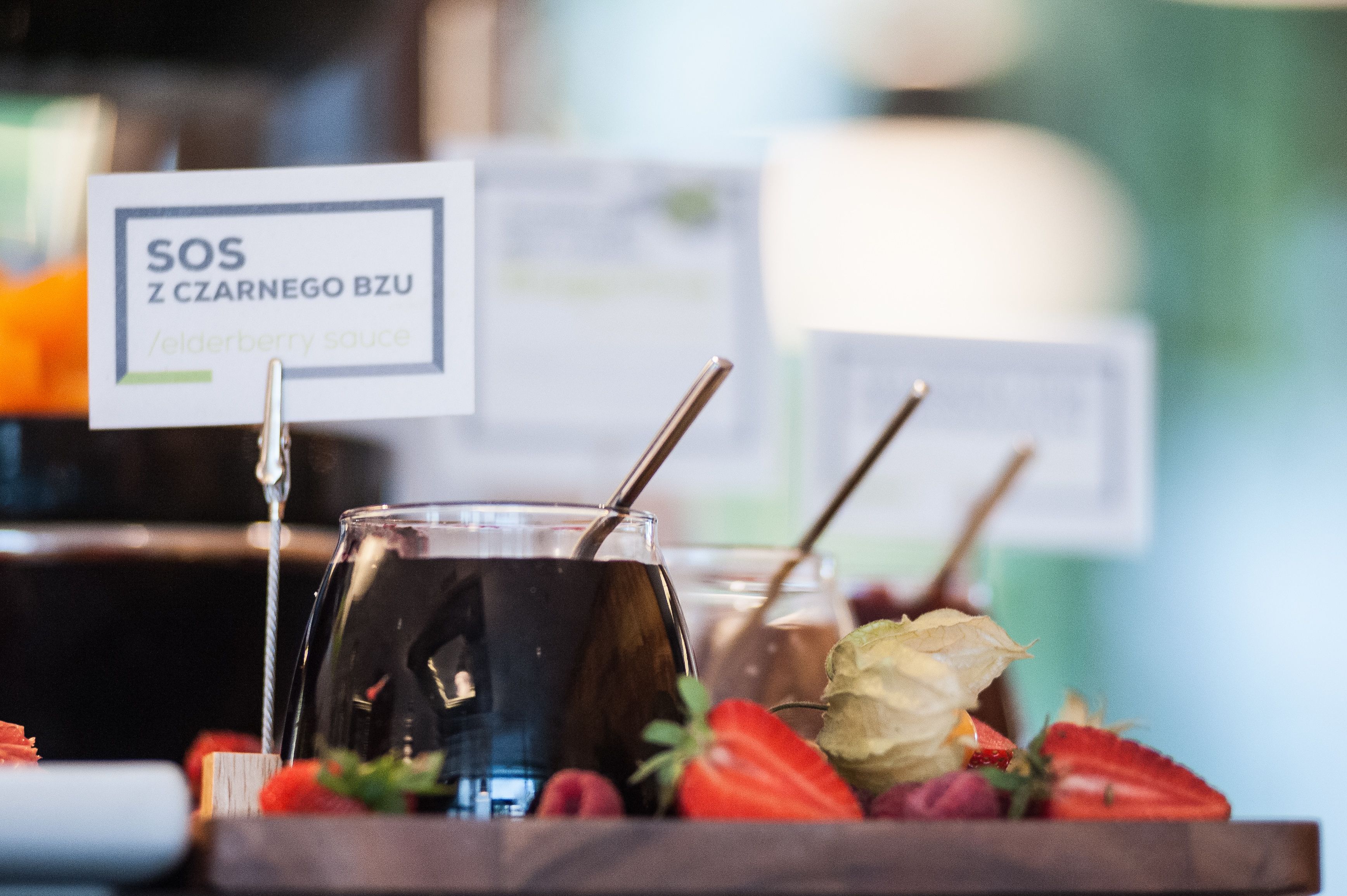 A glass of dark beverage with a straw, accompanied by strawberries and a label reading "SOS Z CZARNEGO BZU / elderberry sauce".