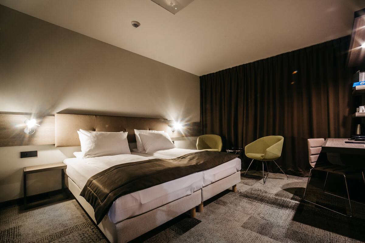 Ein modernes Hotelzimmer mit einem großen Bett, zwei Nachttischen, einem Schreibtisch mit Stuhl und dunklem Vorhang im Hintergrund.