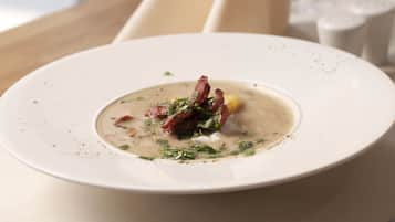 Un plato blanco con sopa cremosa adornada con hierbas y tocino, sobre una mesa color beige.