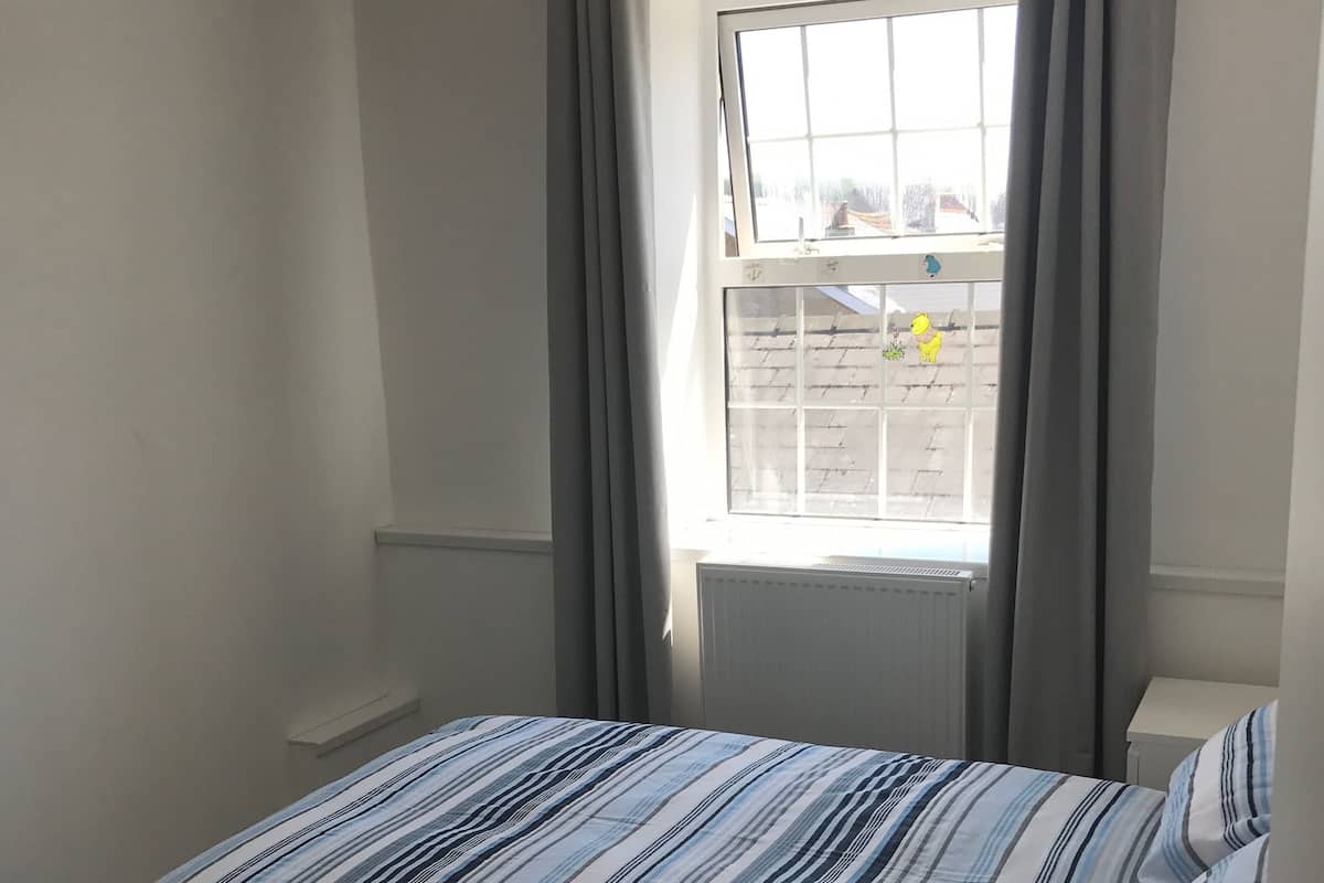 A single bed with a striped bedspread, a bedside table, a radiator, and a window with curtains.