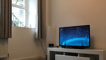 A room with a TV on a white stand, a window with curtains, and a radiator.