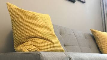 Un sofà gris amb dos coixins grocs, una paret amb tres quadres emmarcados i una finestra amb cortines.