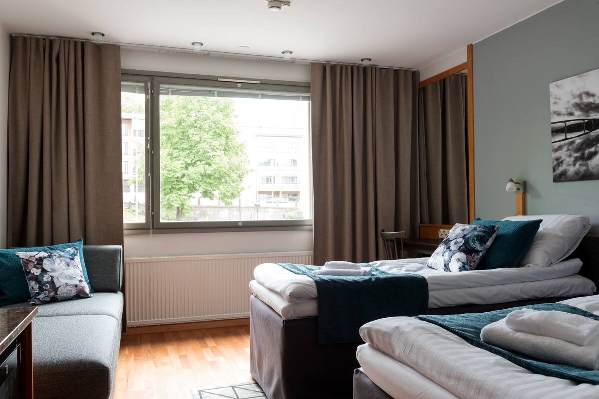 A hotel room with a bed, a sofa, a window with curtains, a radiator, and a wall-mounted artwork.