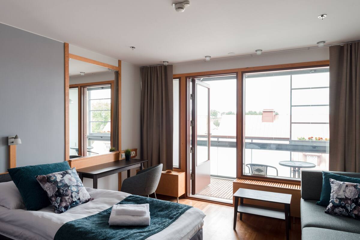 A hotel room with a bed, a desk, a chair, a bench, and a balcony with a table and chairs.