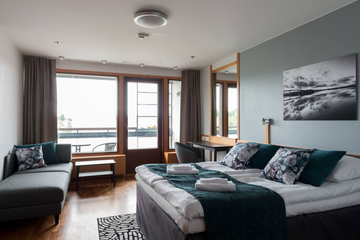 A modern hotel room with a bed, a sofa, a desk, and a view of the outdoors.