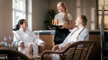 Three people in a spa setting, two in white robes and one serving drinks.