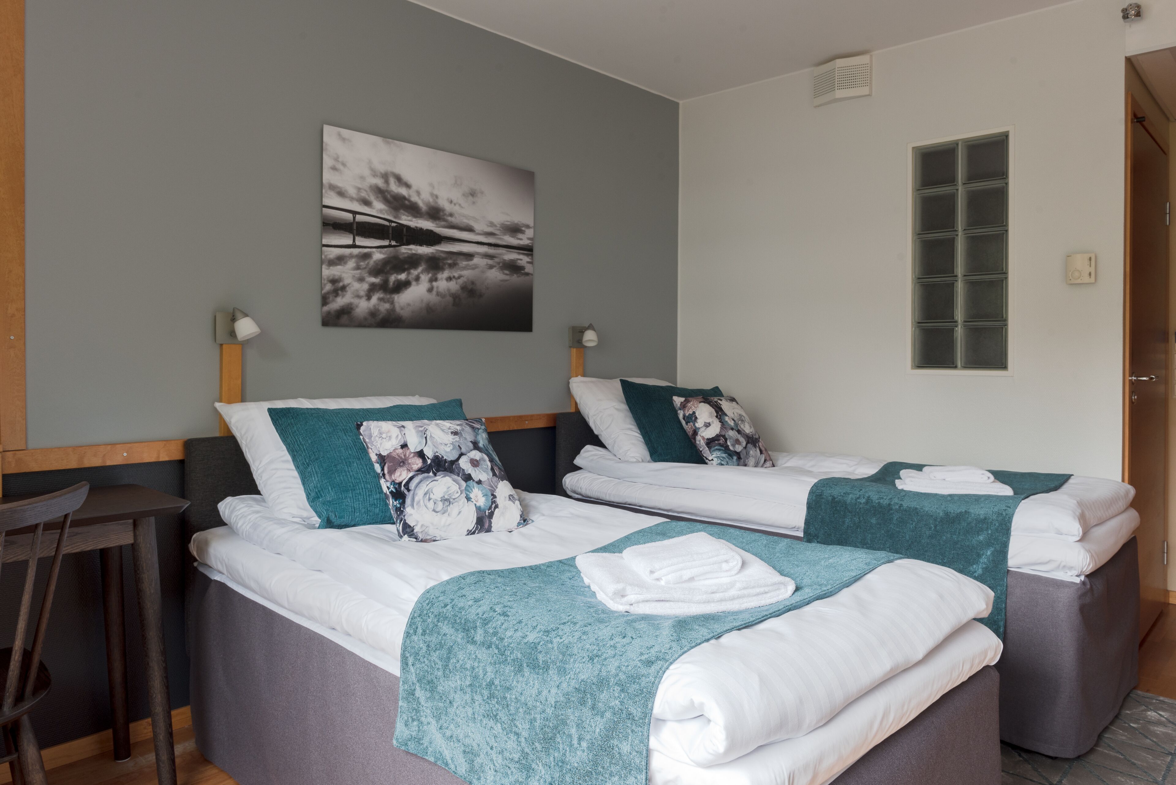 Une chambre avec deux lits, chacun recouvert d’une couverture turquoise et agrémenté d’oreillers à motifs floraux. On y trouve un bureau avec une chaise, une lampe fixée au mur et une fenêtre donnant sur un paysage agréable.