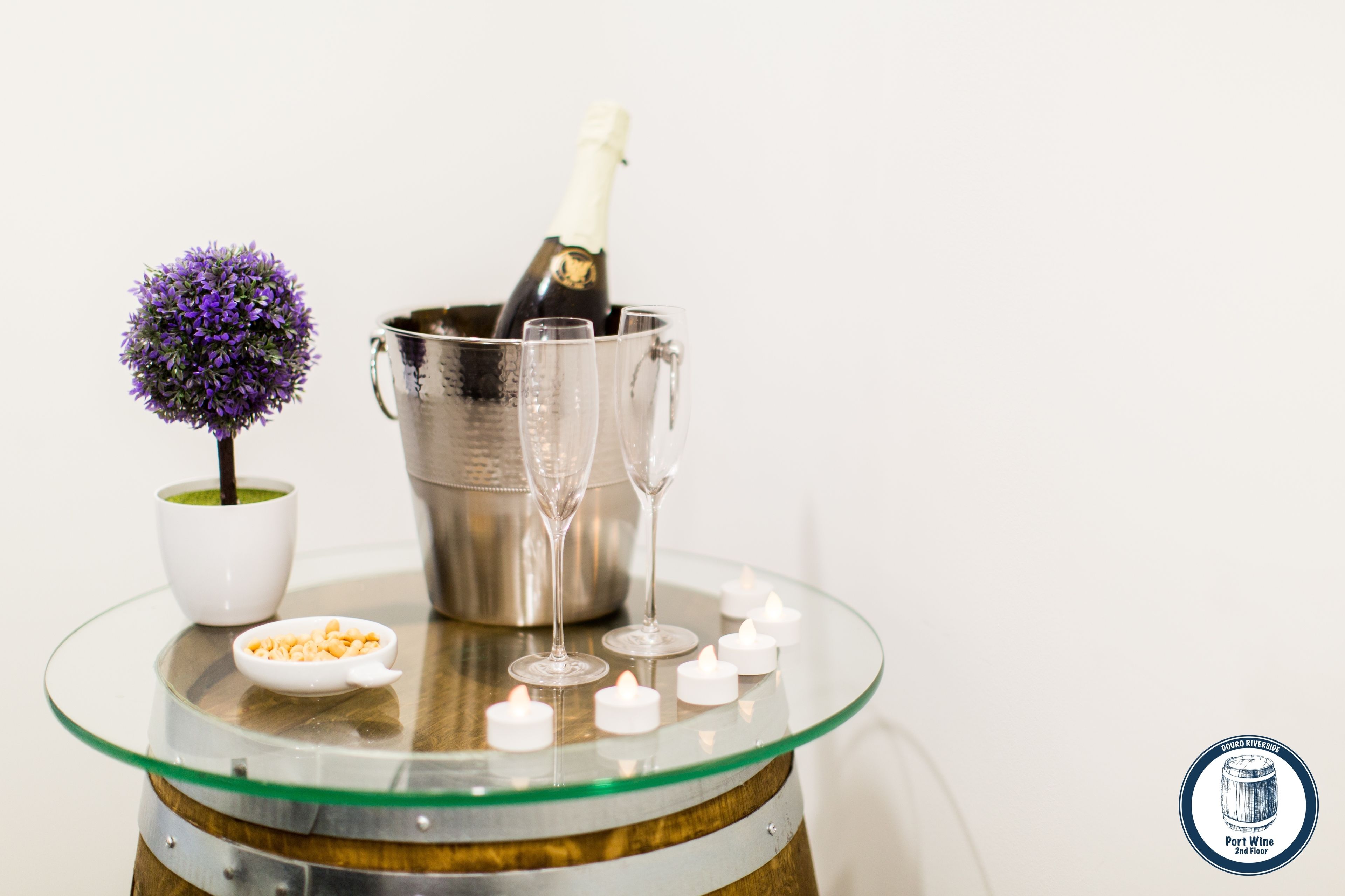 A glass table with a champagne bottle, two glasses, a small bowl of nuts, a white cup, and a decorative purple flower.