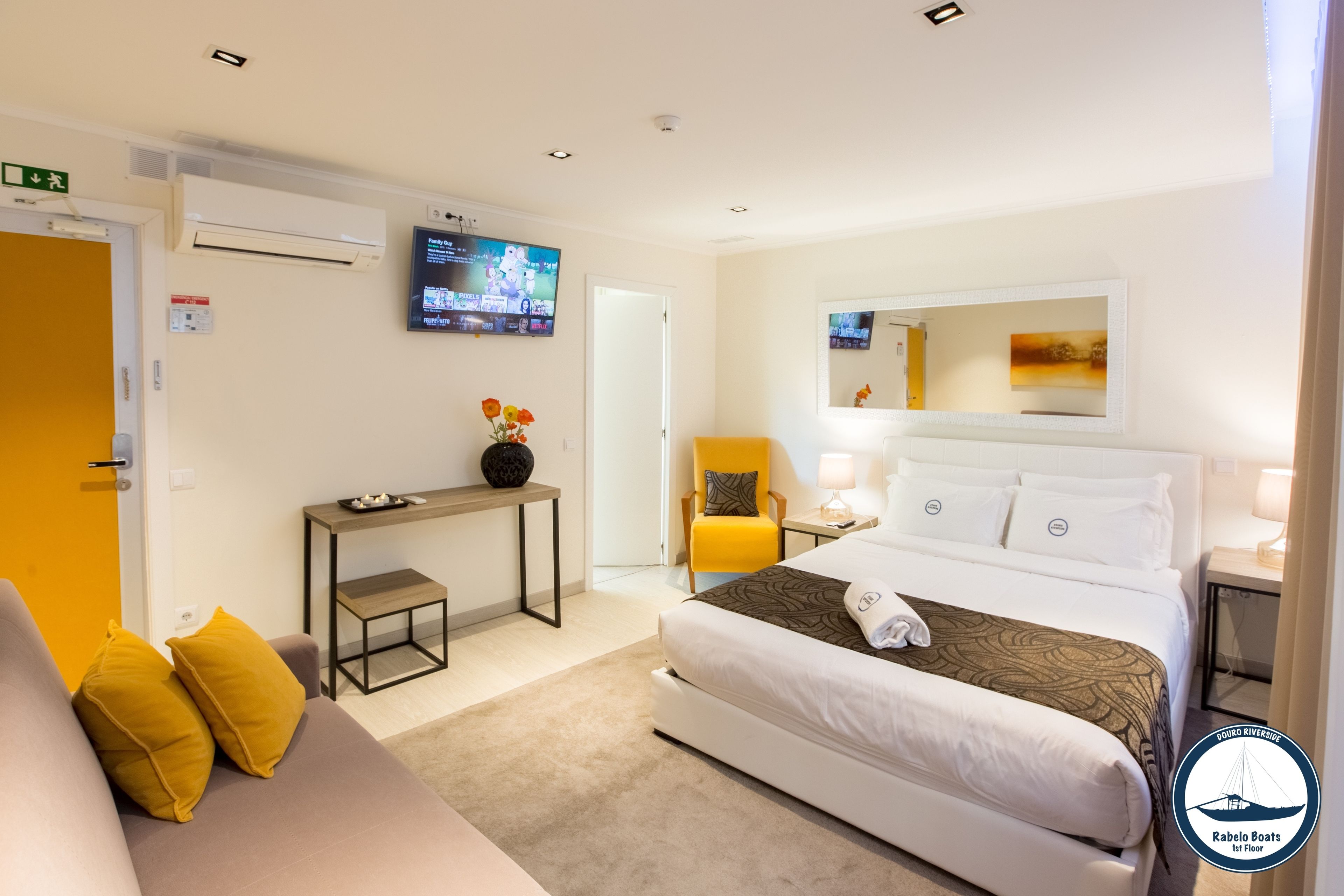 Une chambre d’hôtel avec un lit, un canapé, une petite table sur laquelle est posé un vase de fleurs, une télévision et un miroir au mur.