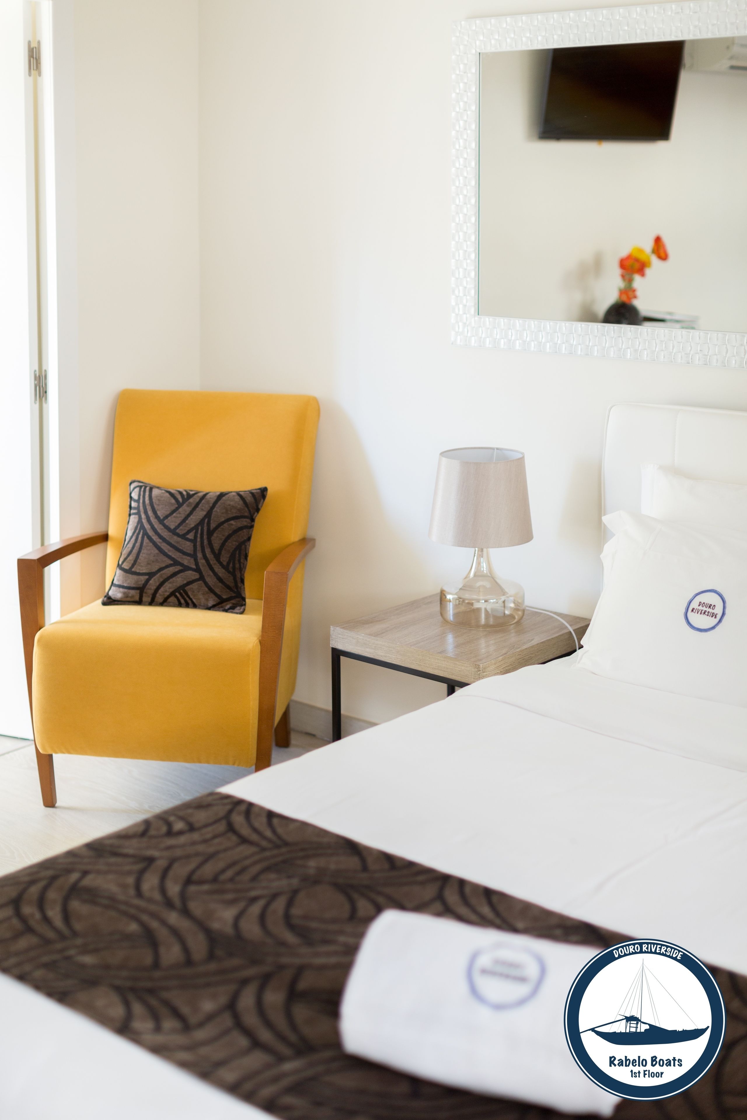 Habitación de hotel con cama, una silla con almohada estampada, una mesita de noche con lámpara y un espejo en la pared.