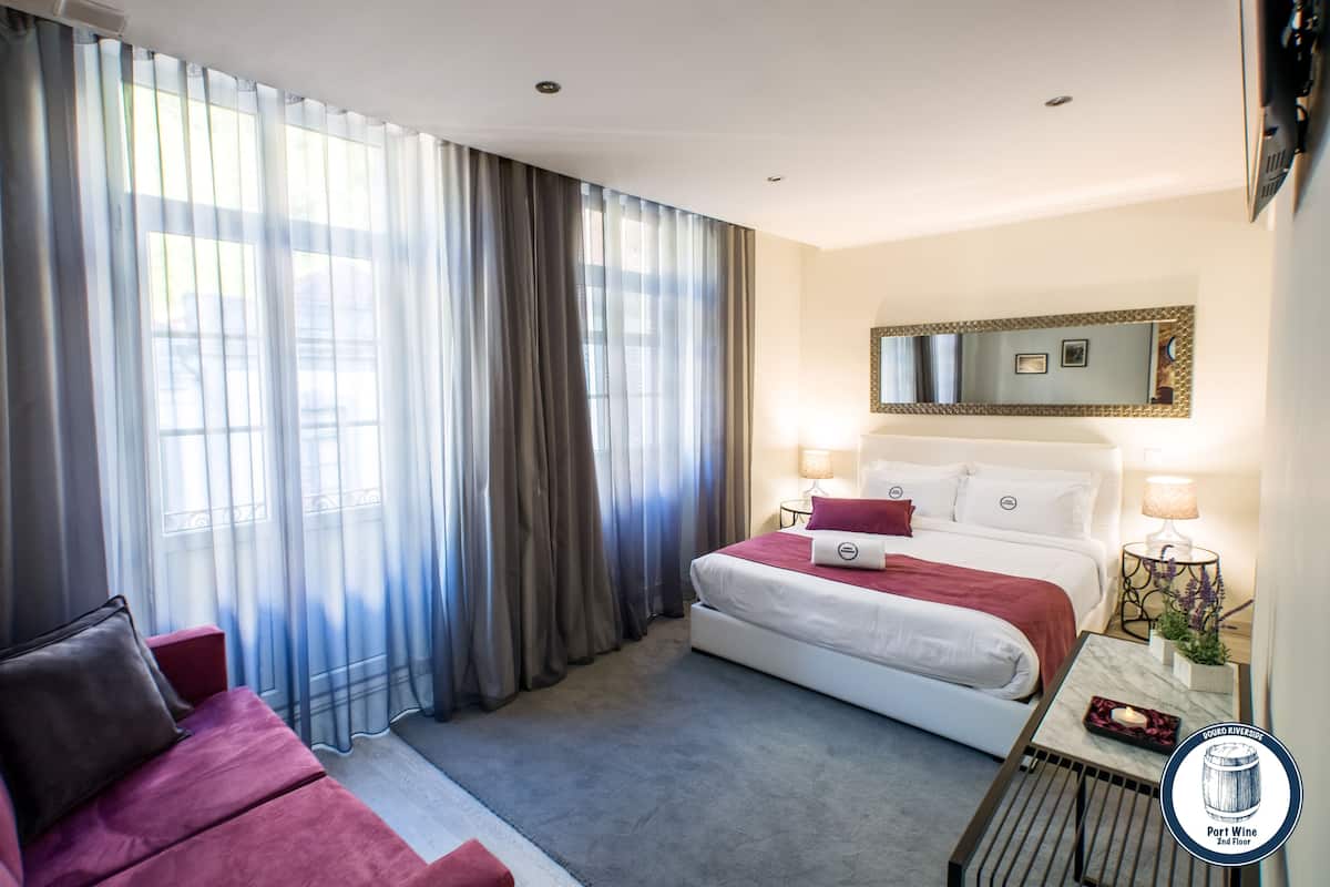 A hotel room with a bed, a sofa, a nightstand with a lamp, and a large window with curtains.