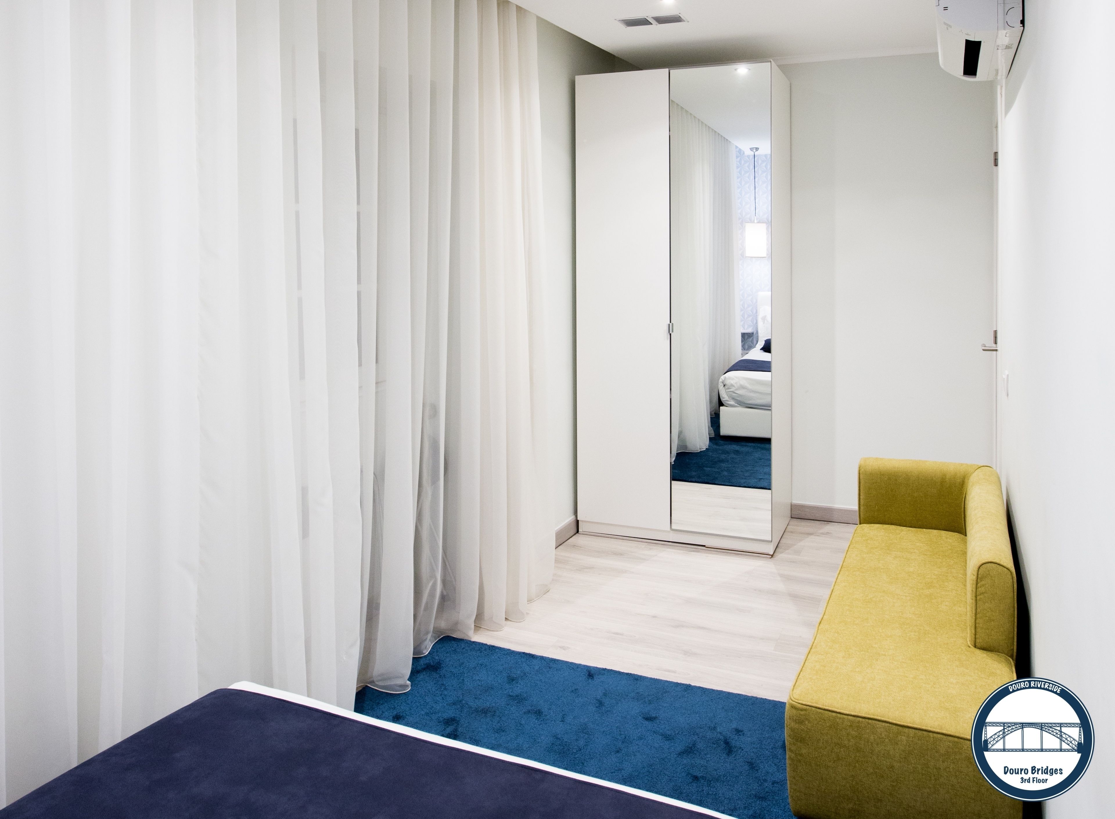 Hotellihuone, jossa on sänky, keltainen nojatuoli, sininen matto ja suuri peili, johon heijastuu kylpyhuone valkoisella pesualtaalla ja sinisillä verhoilla.