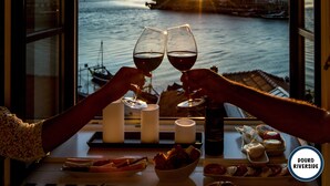 A table with a bottle of wine, glasses, candles, and food. A view of a river and buildings is visible through a window. - Douro Riverside Apartments (Vila Nova de Gaia)