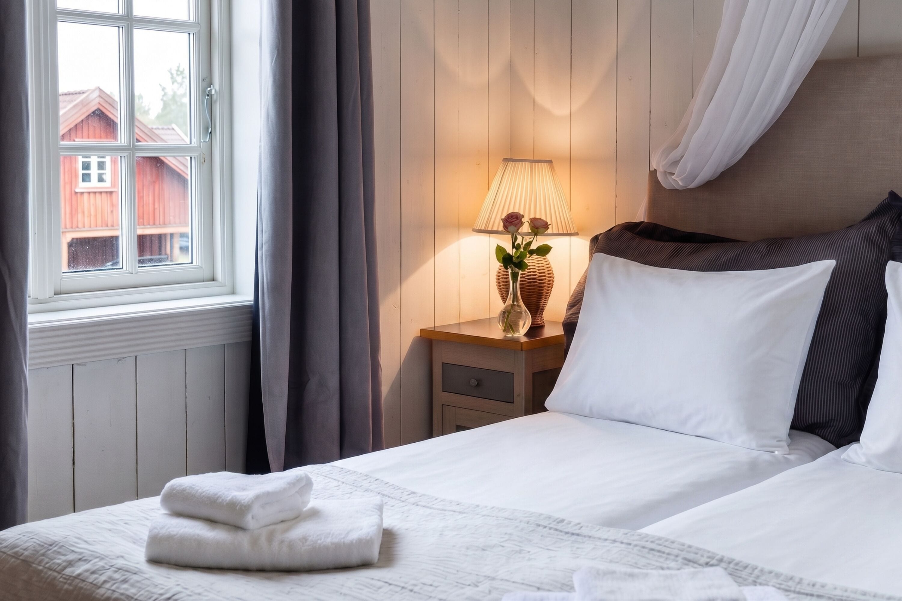 Une chambre d’hôtel avec un lit, une table de chevet avec une lampe et un vase, une fenêtre avec des rideaux et une vue sur un bâtiment rouge à l’extérieur.