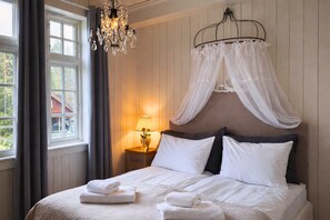 A bedroom with a bed, white curtains, a chandelier, and a nightstand with a lamp. - Eggedal Borgerstue (Sigdal)