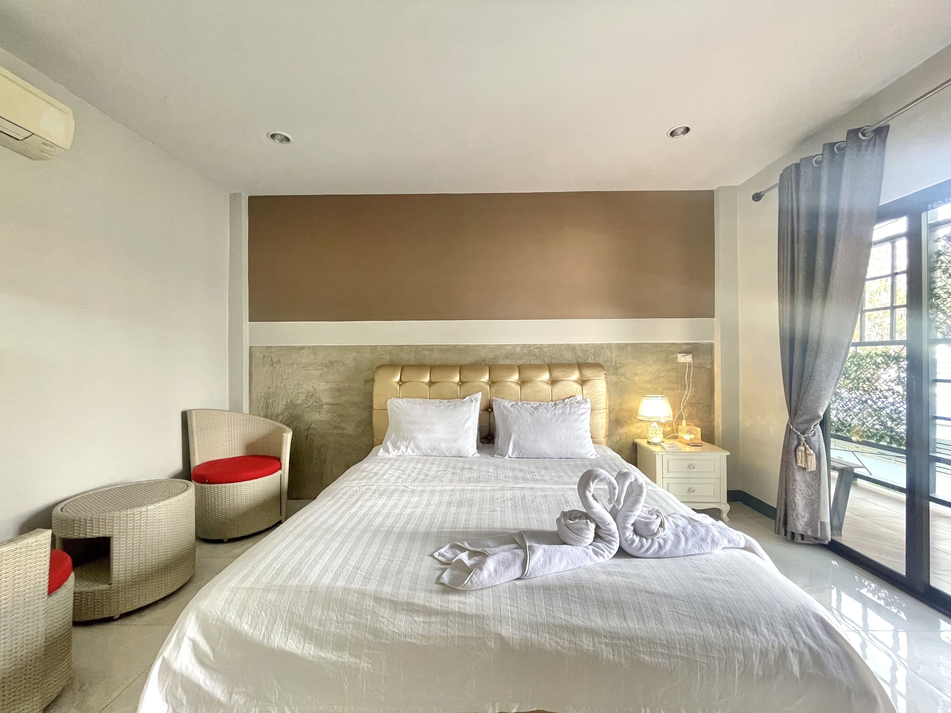 A hotel room with a large bed, a headboard, a nightstand with a lamp, a wicker chair with a red cushion, and a view of greenery through the window.