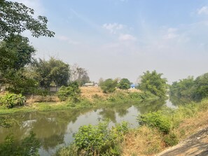 A calm river with greenery and a few buildings in the background. - Smaids Room (Lampang)