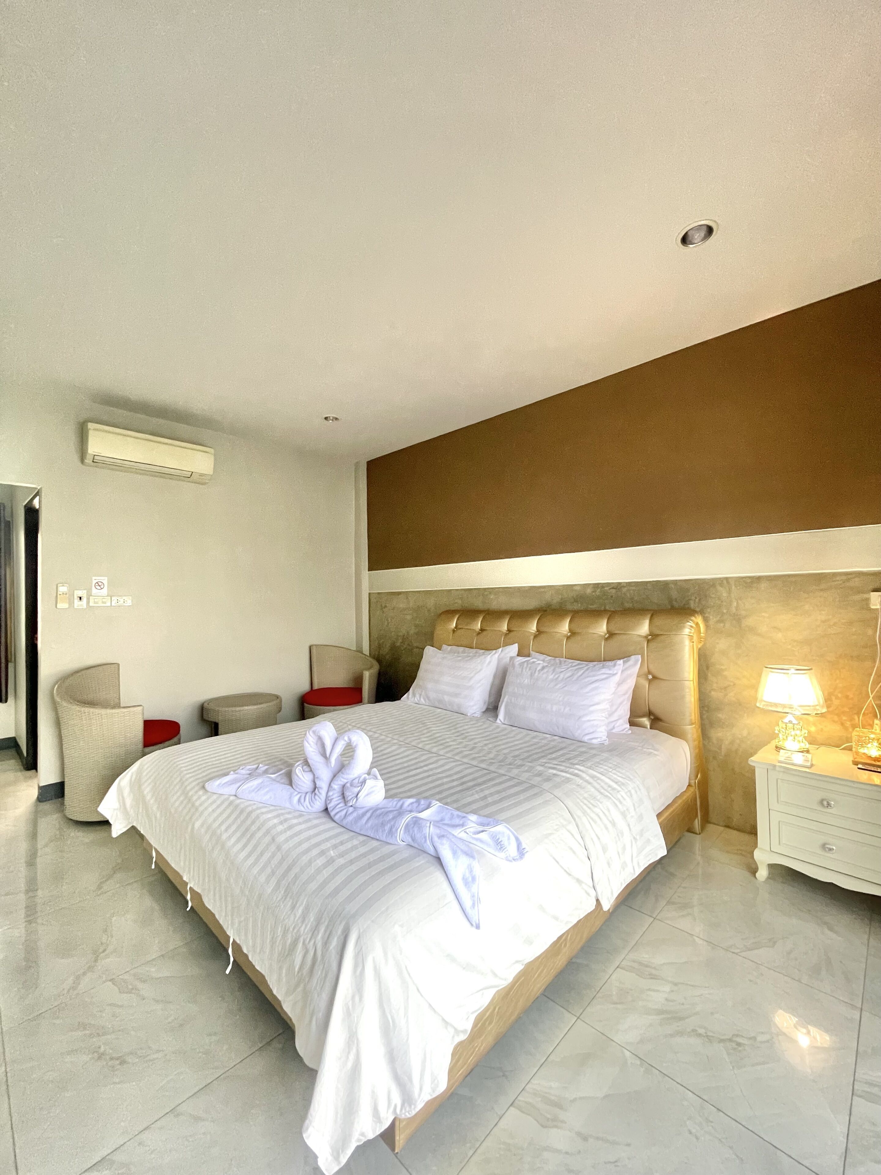 Una habitación de hotel con una cama grande, un cabecero, una mesita de noche con lámpara, una silla, una pequeña mesa y un equipo de aire acondicionado.