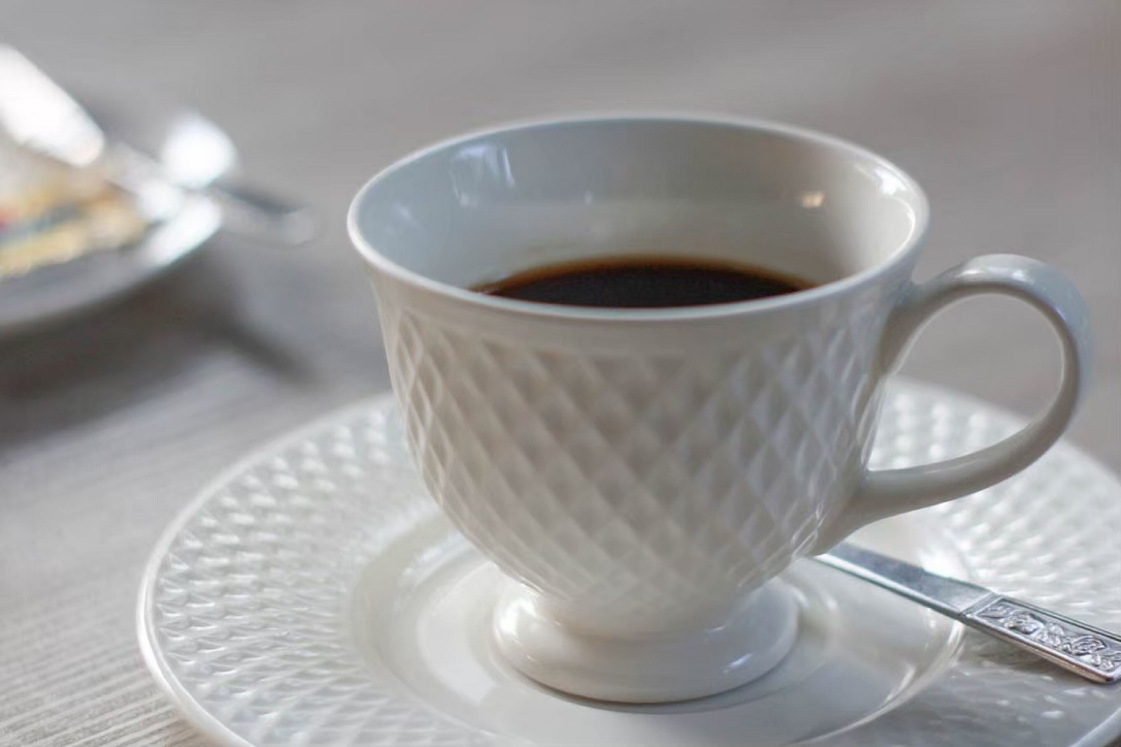 Una taza de cerámica blanca con un diseño texturizado, llena de un líquido oscuro, probablemente café, sobre un platillo y cuchara a juego.