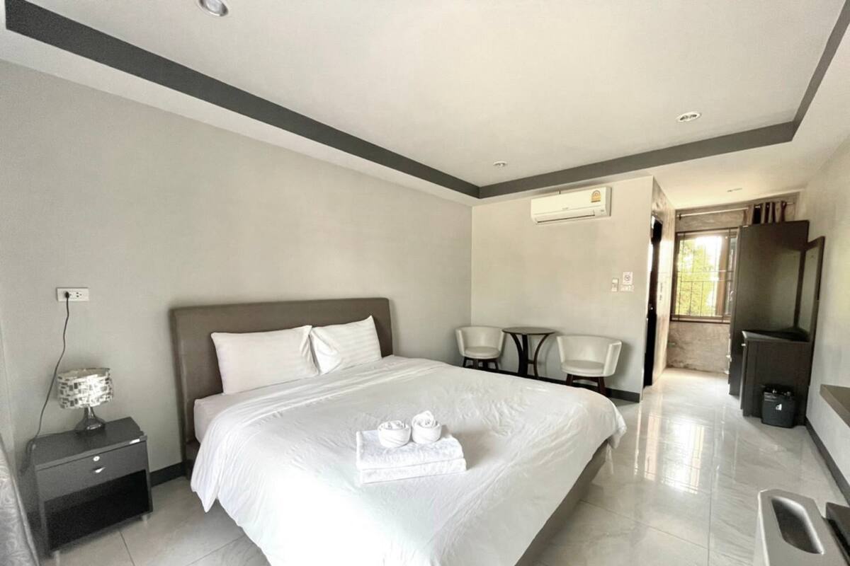 A modern hotel room with a large bed, a bedside table, a lamp, two chairs, a small table, a TV, and an air conditioning unit.