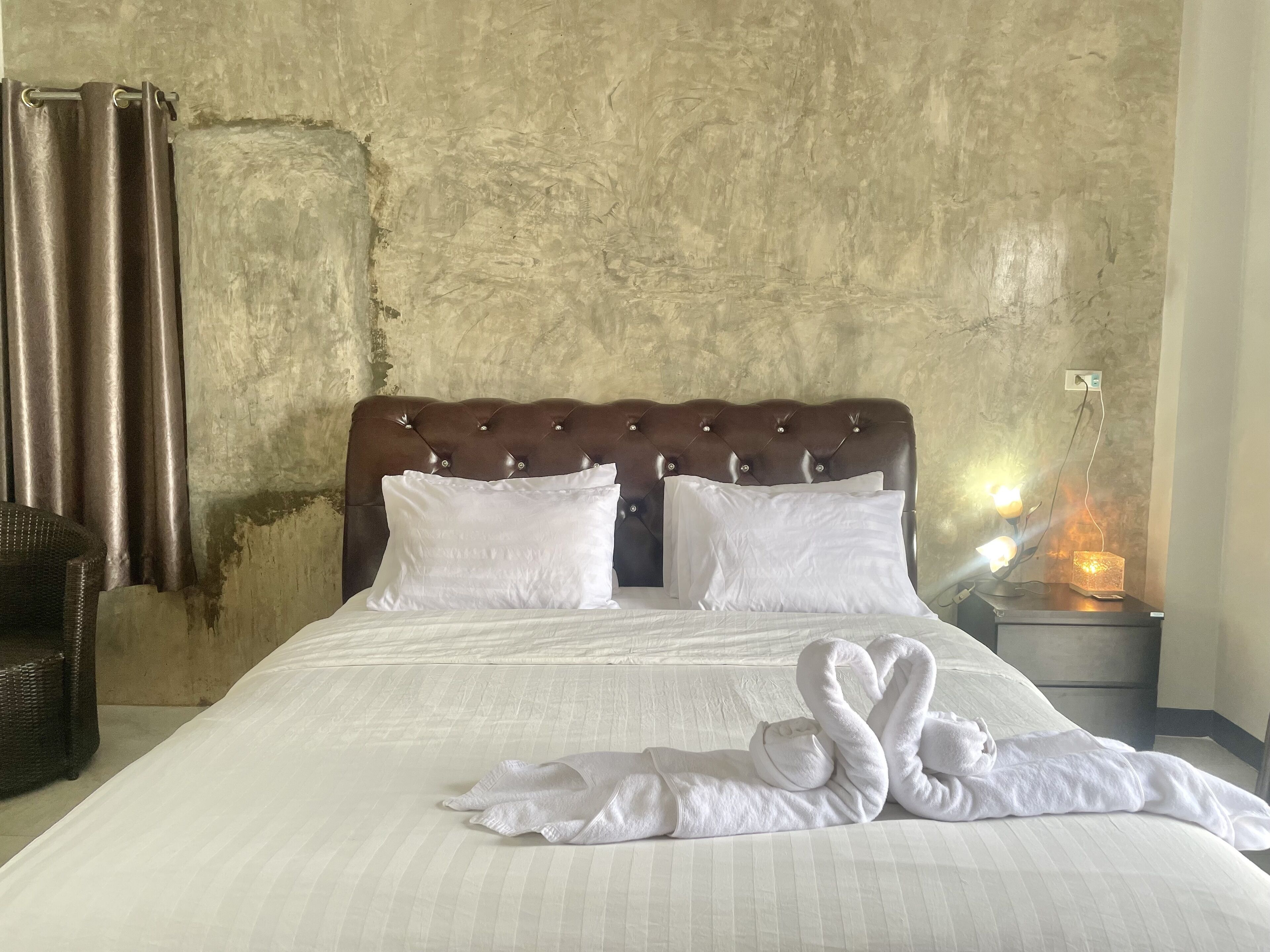 A neatly made bed with white linens, a brown tufted headboard, and two pillows. There is a wicker chair to the left and a bedside table with a lamp and a decorative item.