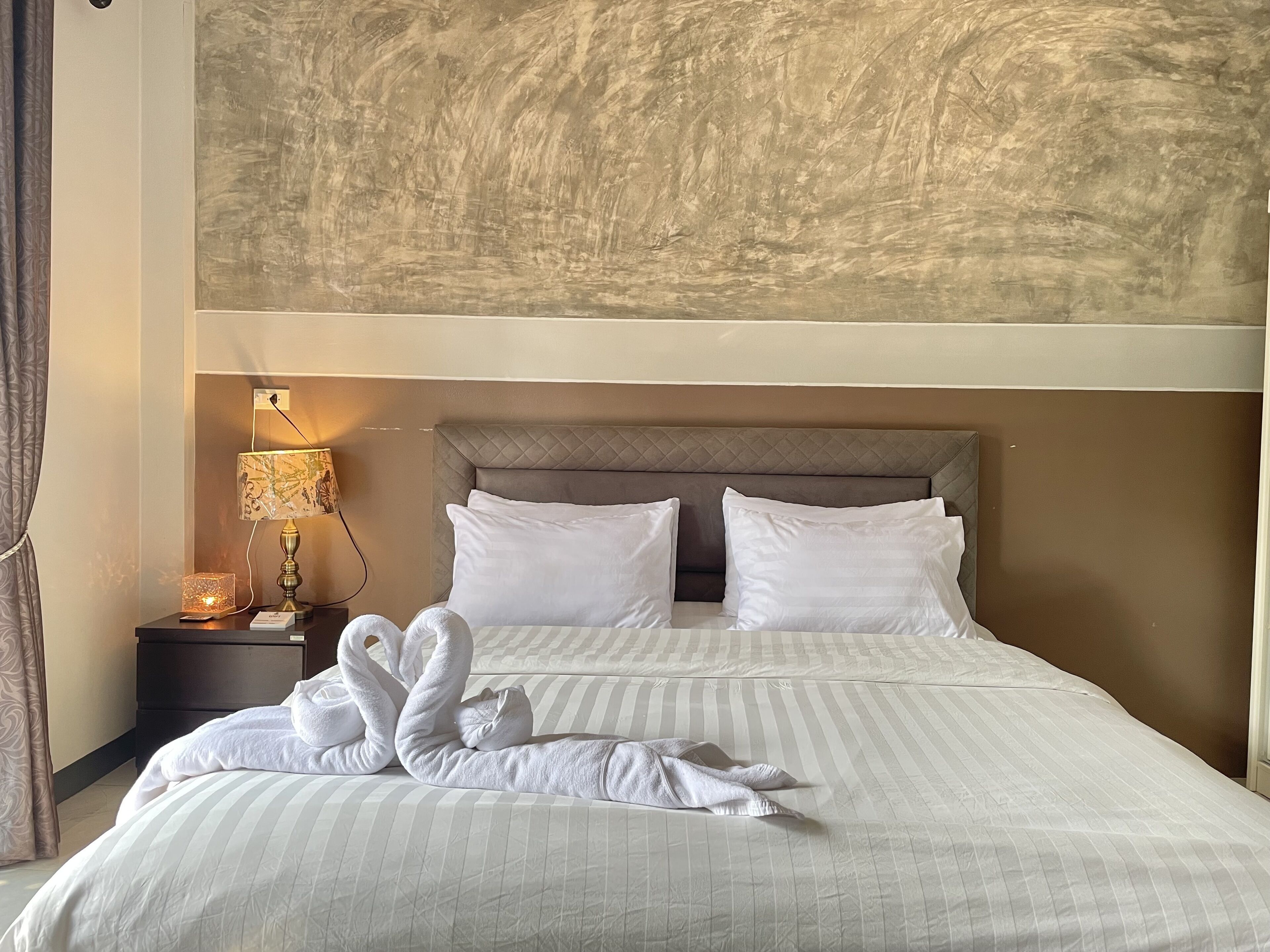 Habitación de hotel con una cama grande, dos almohadas, una mesita de noche con lámpara y una pared con textura.