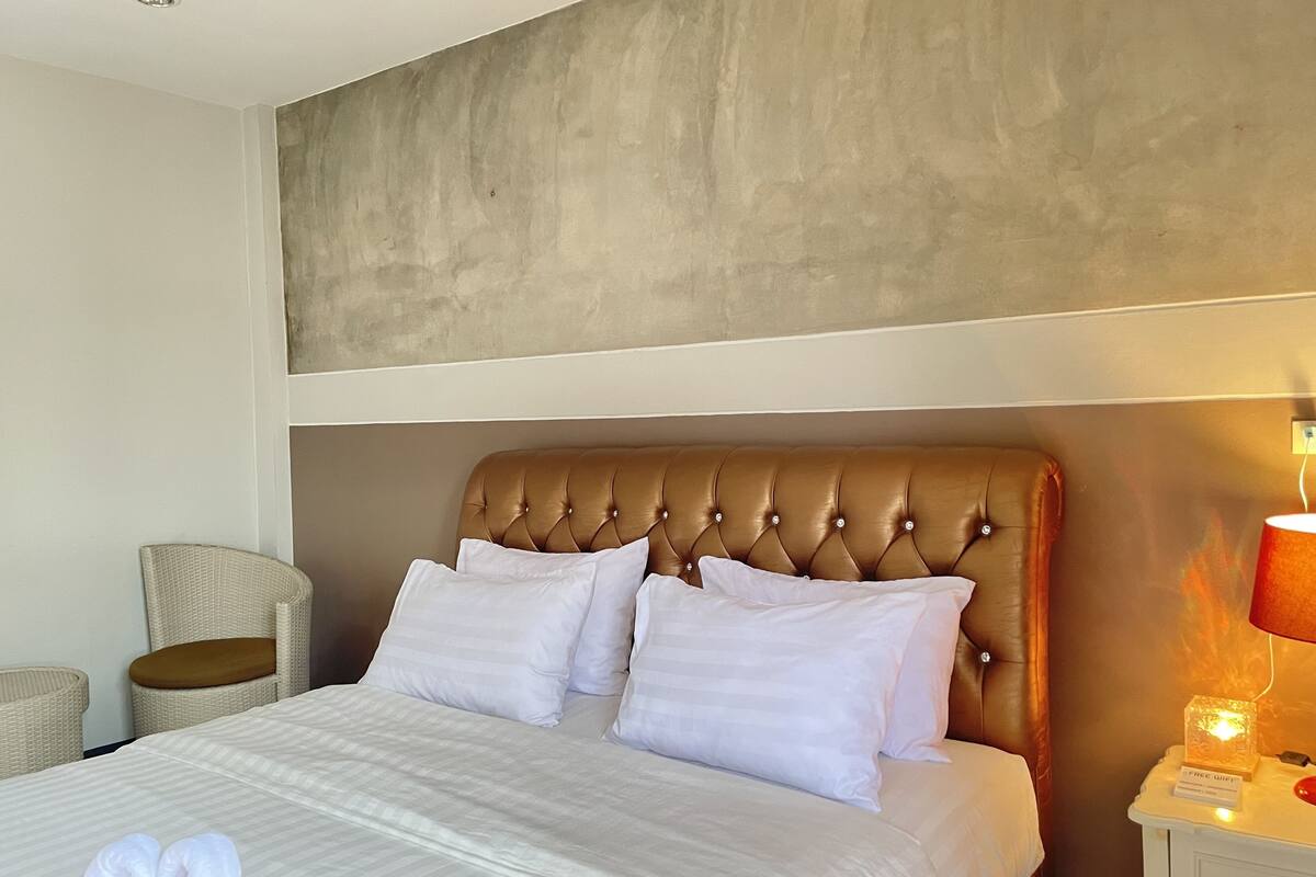 A hotel room with a large bed, a tufted headboard, white bedding, a wicker chair, and a nightstand with a lamp and a sign indicating free WiFi.