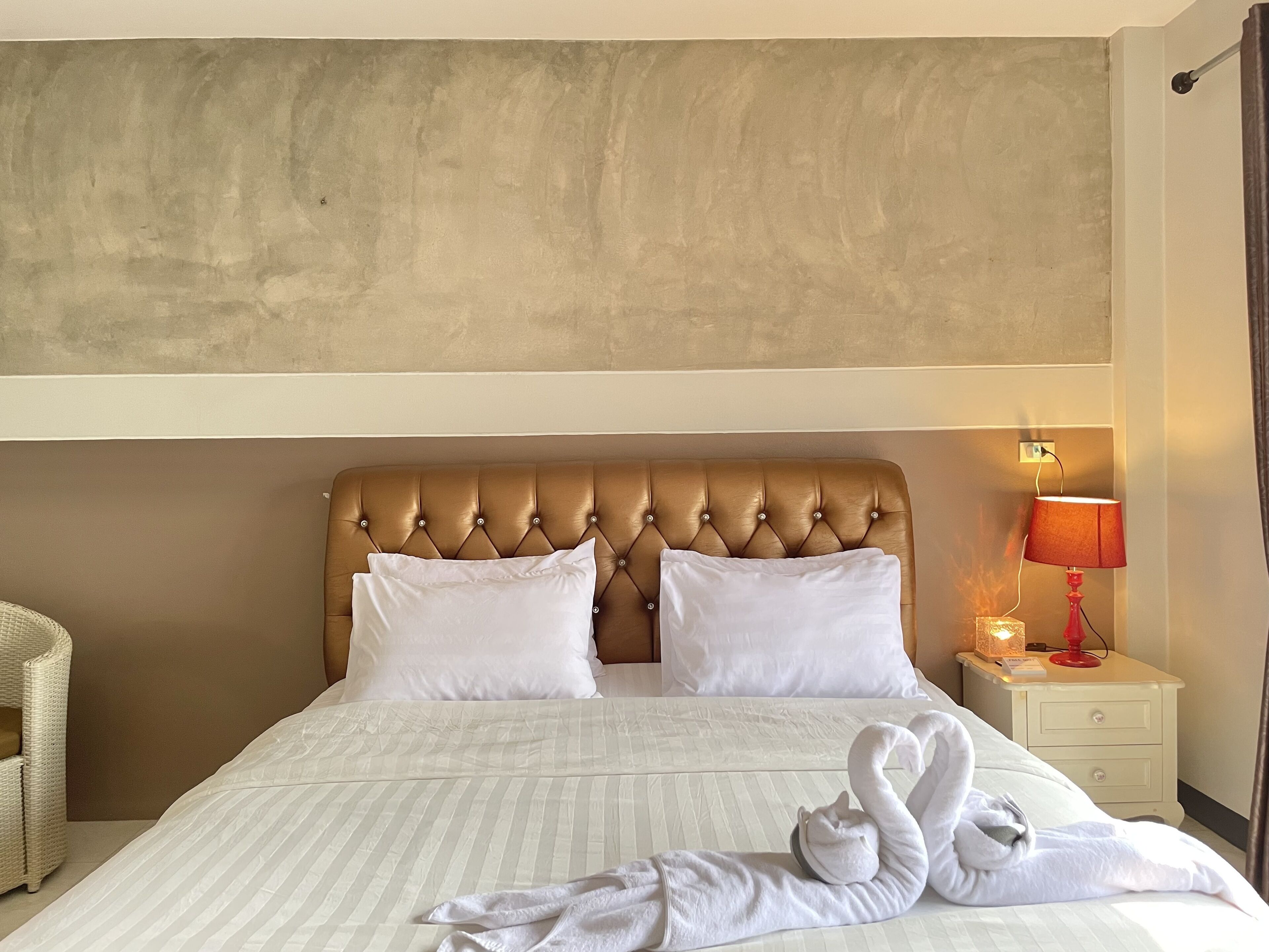 A hotel room with a tufted headboard, two white pillows, a bed with striped bedding, a red lamp on a bedside table, and a swan-shaped towel arrangement.
