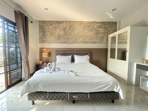 A large bed with a quilted headboard, a bedside table with a lamp, and a view of the outdoors through a window with curtains. - Smaids Room (Lampang)