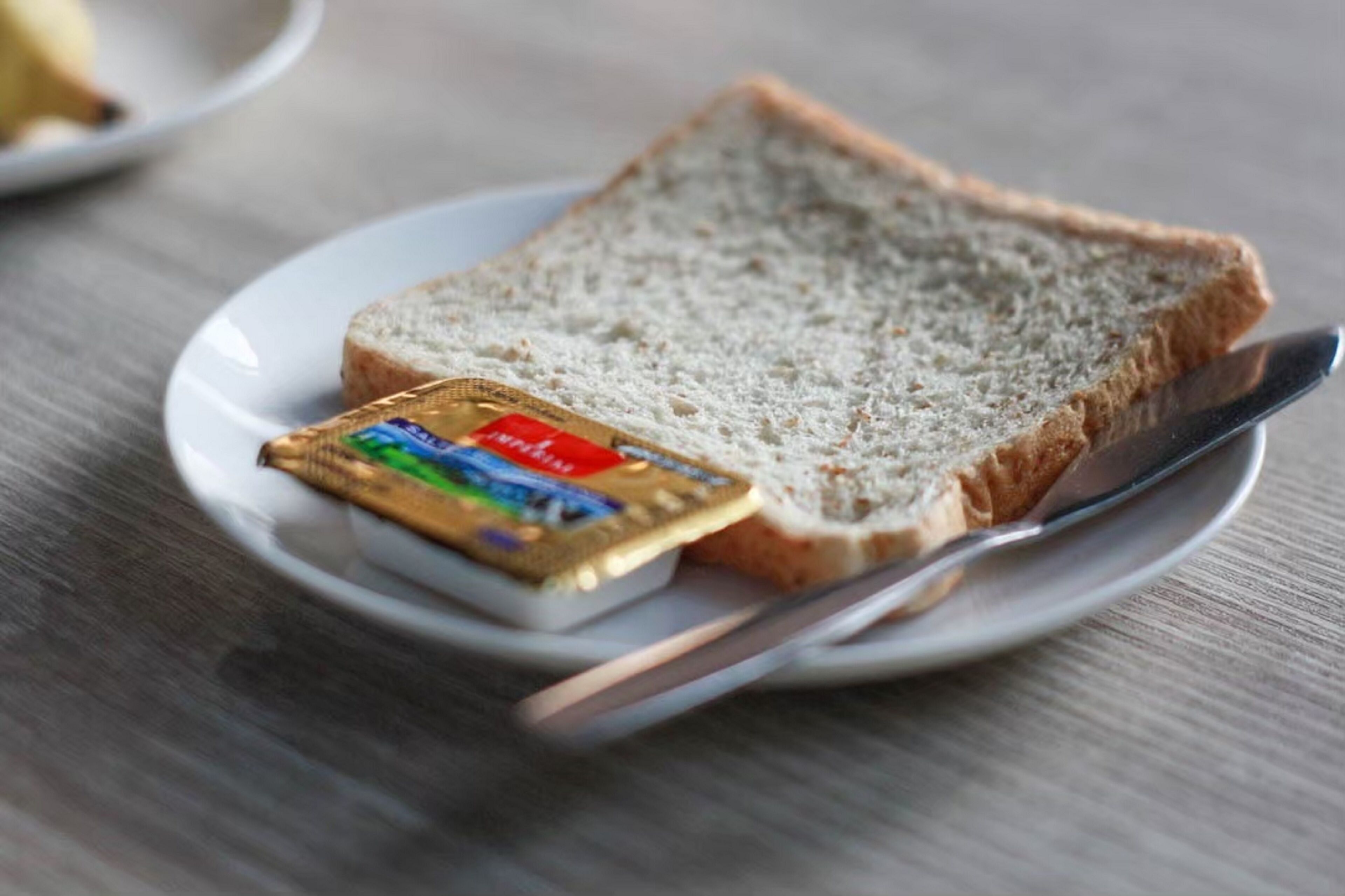 A slice of white bread on a white plate with a butter knife and a packet of butter.
