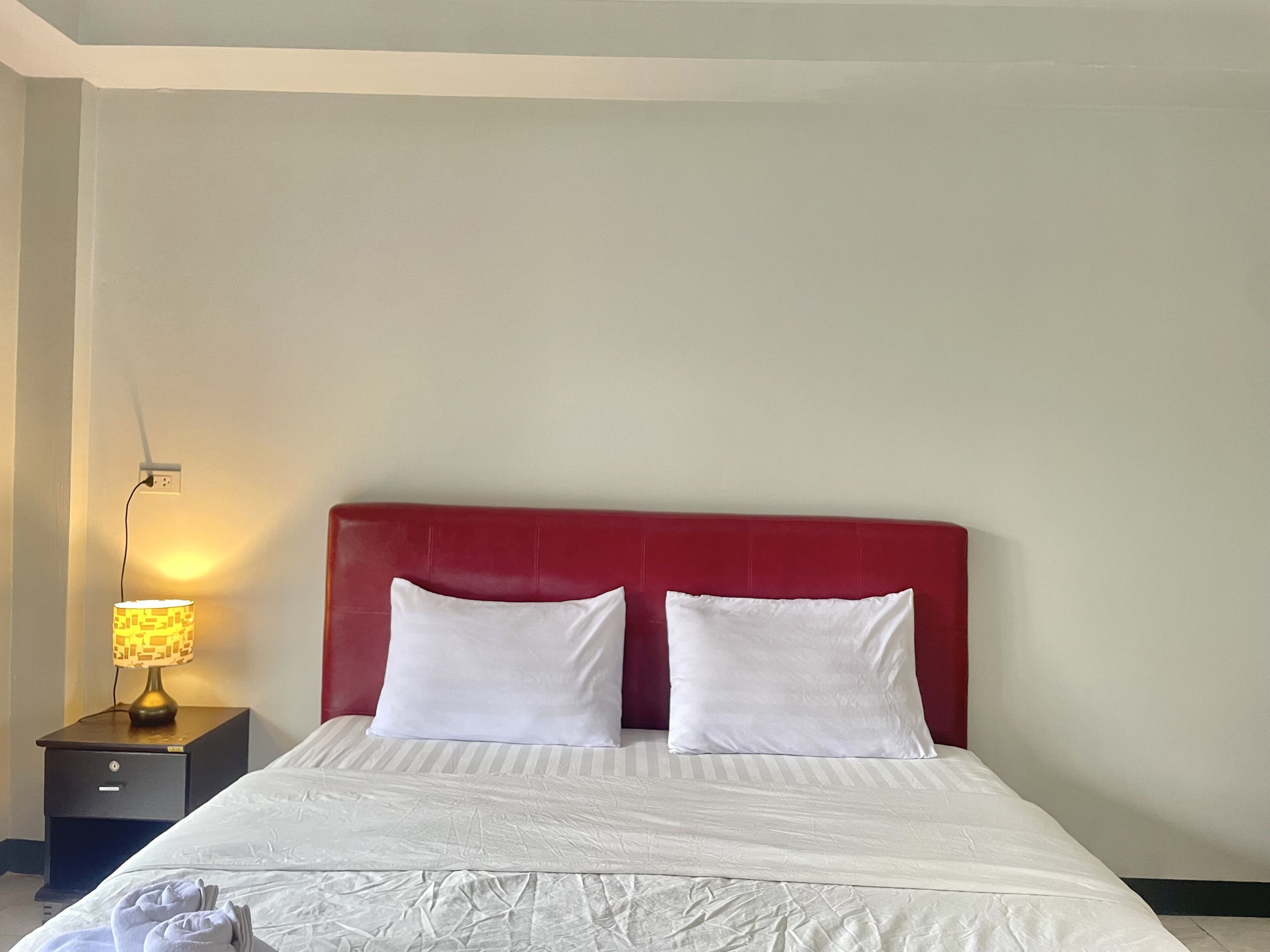 Cama bem arrumada com lençóis brancos e duas almofadas, cabeceira vermelha e abajur com cúpula estampada.