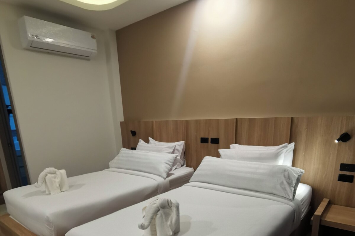 Une chambre d’hôtel avec deux lits, chacun équipé de draps blancs et d’une serviette. On y trouve un climatiseur mural, des têtes de lit en bois et une petite table de chevet en bois avec une lampe.