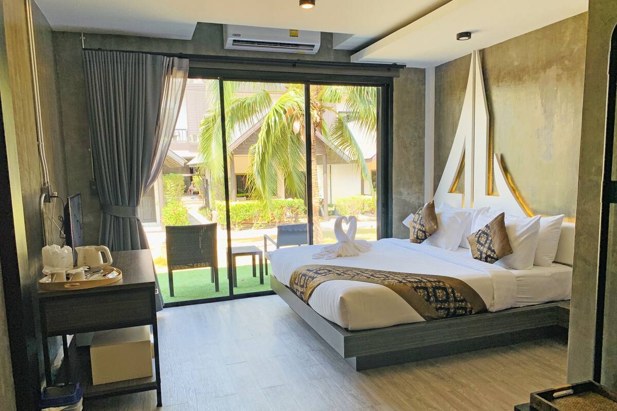Ein modernes Hotelzimmer mit einem großen Bett, einem Schreibtisch mit Frühstücksgebäck, einem Stuhl und Blick auf einen Innenhof mit Palmen.