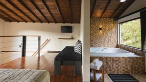 A bedroom with a wooden floor, a bed, a bench, a small table with towels, a TV on the wall, and a window with a view of greenery. - Pousada Ilha da Magia (Florianópolis)