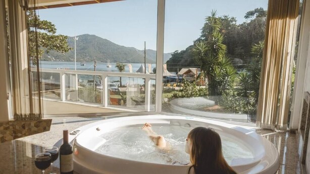 A person relaxing in a hot tub with a scenic view of a lake and mountains. - Pousada Ilha da Magia (Florianópolis)