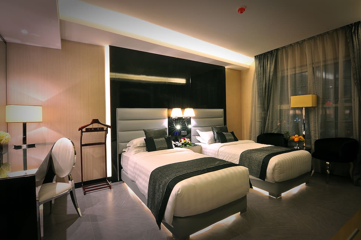 A hotel room with two beds, a desk with a chair, a bedside table with lamps, and a large window with curtains.