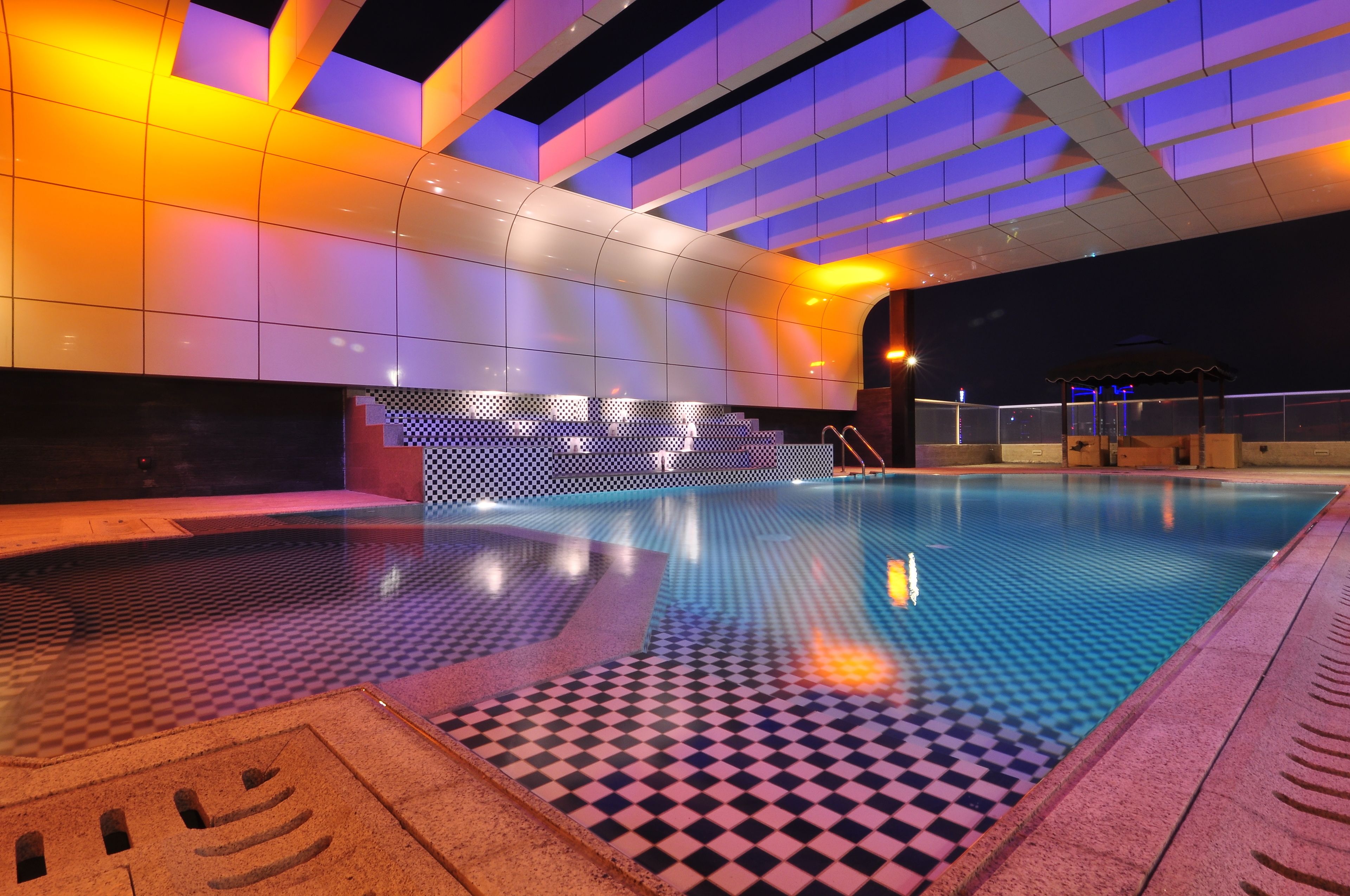 Une piscine intérieure avec des carreaux de sol à motifs géométriques, un mur recouvert de carreaux formant un motif géométrique, et un plafond doté de panneaux lumineux.