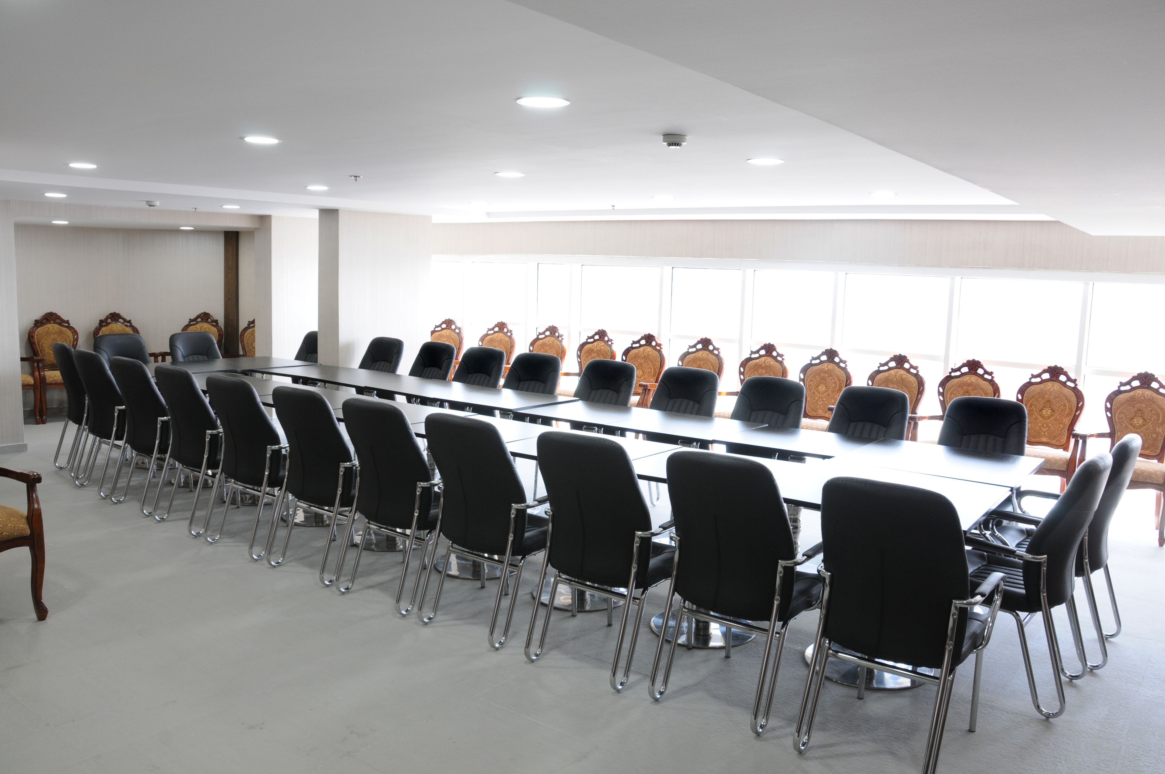 A conference room with a long table and rows of chairs, large windows, and a white ceiling.