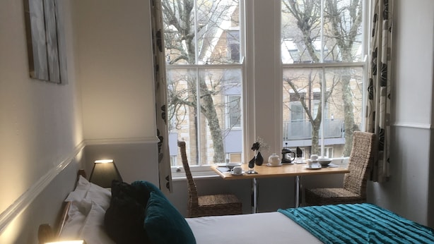 A bedroom with a bed, bedside lamp, and a view of trees through the window. - Garswood Guesthouse (Penzance)