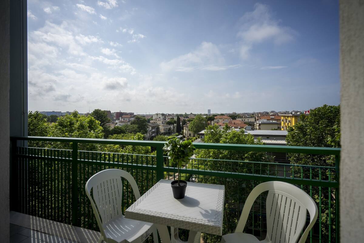 A balcony with a table and two chairs, overlooking a cityscape with buildings and trees.