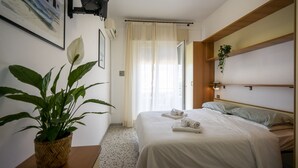 Ein Hotelzimmer mit einem Bett, einer Topfpflanze, einem Fenster mit Vorhängen und einem an der Wand befestigten Regal mit einer Pflanze.