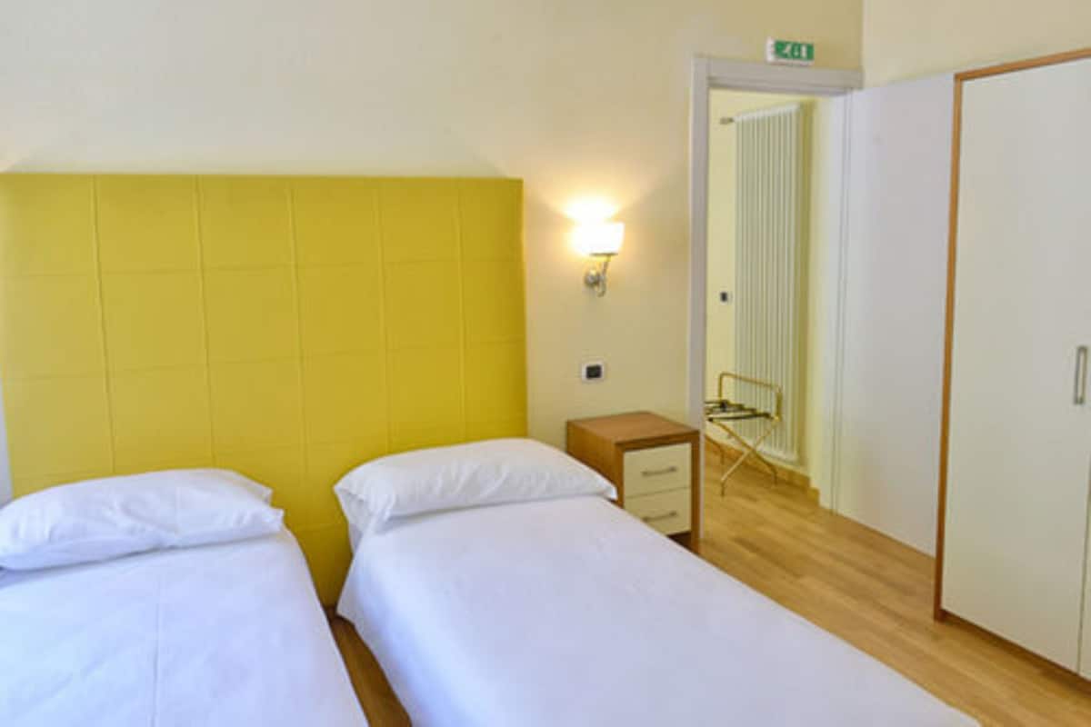 Une chambre avec deux lits, une armoire, une petite table de chevet et une lampe fixée au mur.