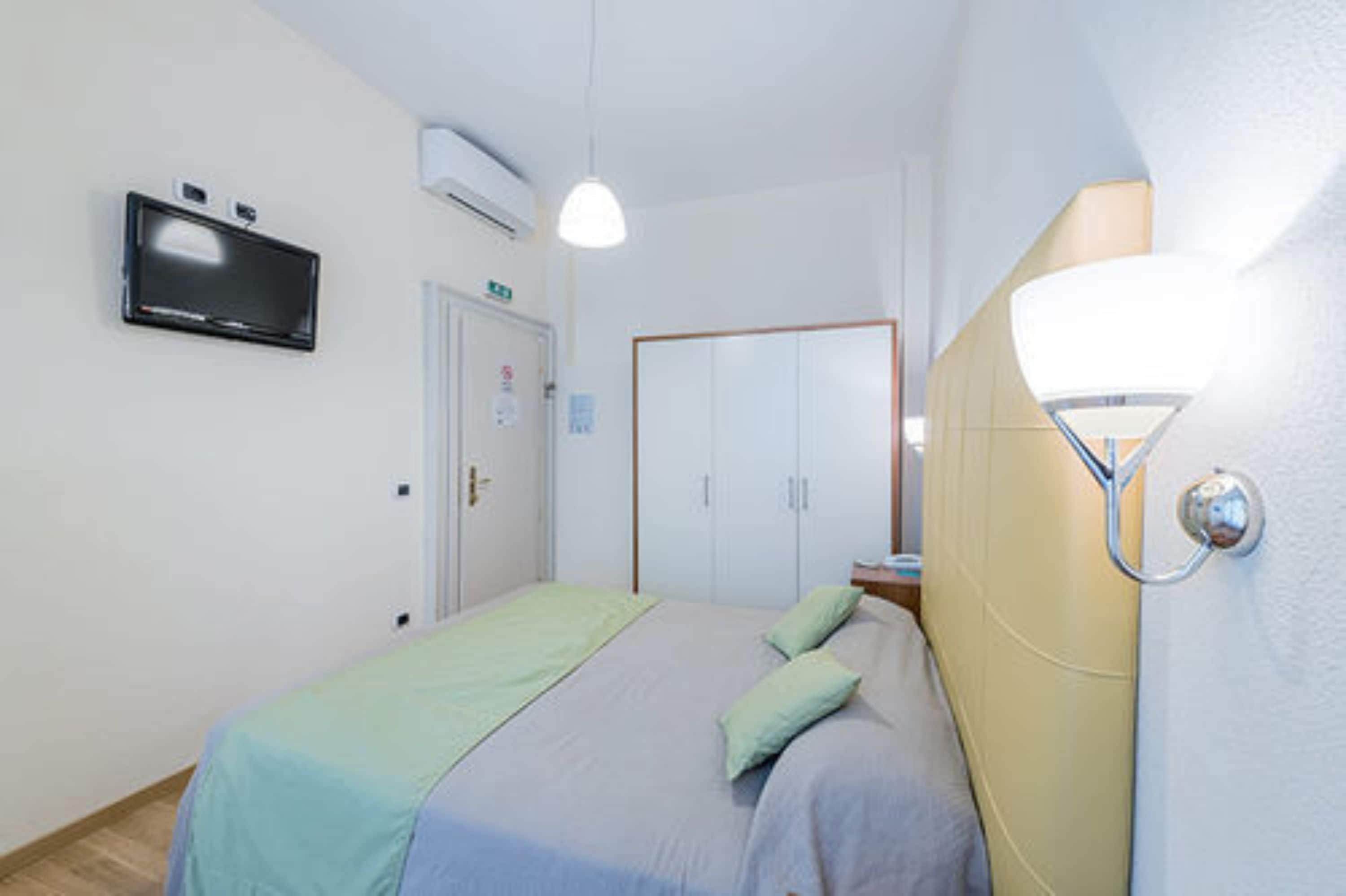 A bedroom with a bed, a television mounted on the wall, a wardrobe, and a bedside lamp.