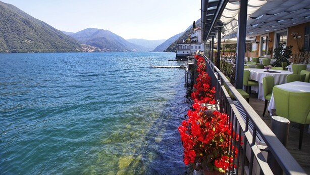 Restaurante frente al lago con terraza al aire libre y vistas a un lago tranquilo y montañas.